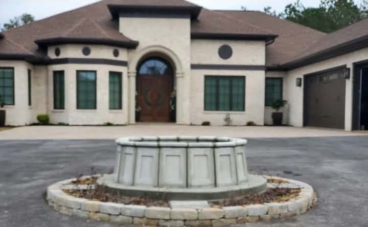 Finished round concrete fountain basin in a driveway circle in front of a stone home, with a stacked stone surround